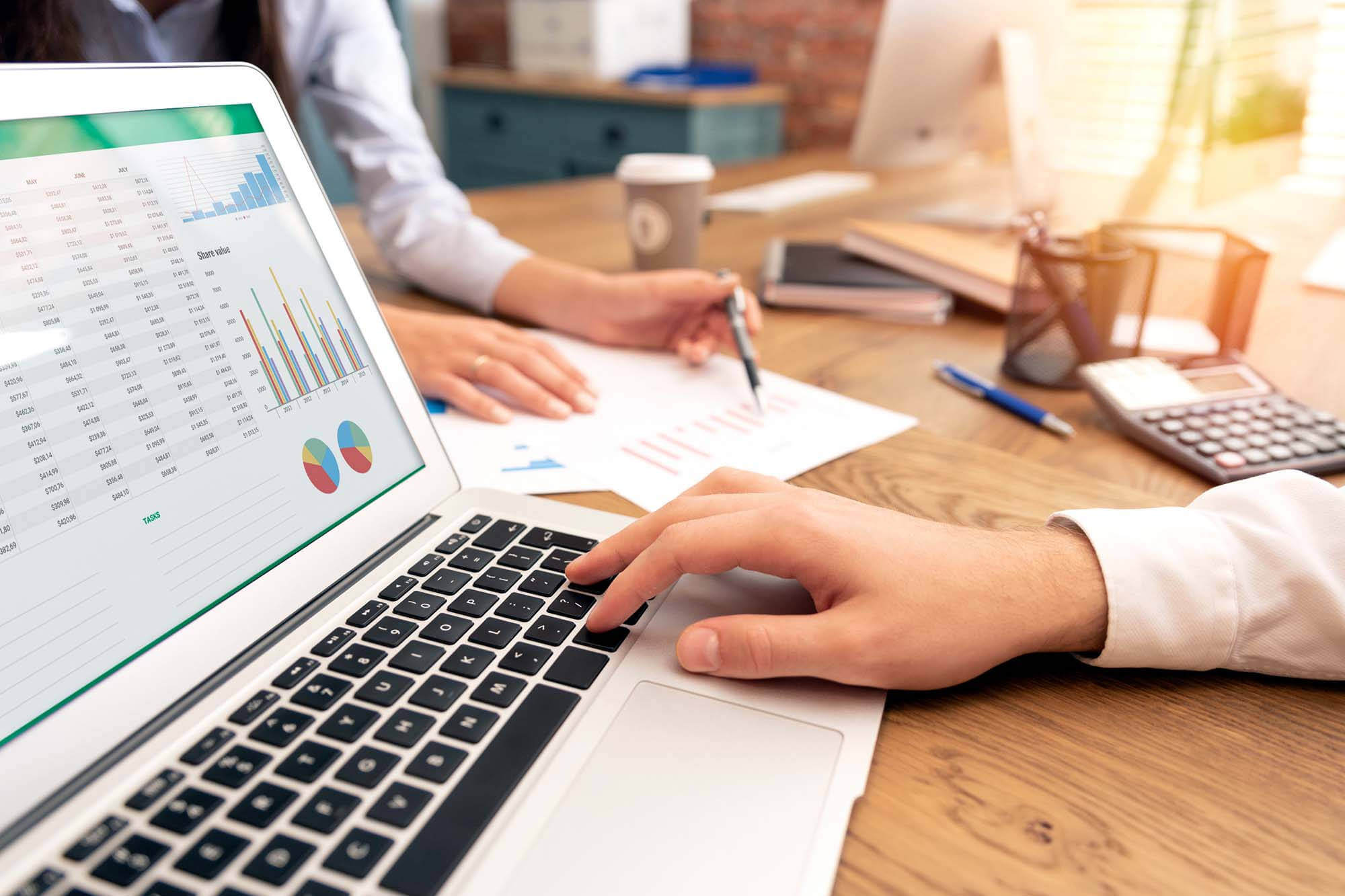 Two colleagues at wooden table reviewing laptop with charts and data